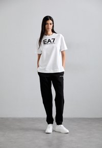 Bijela pamučna majica s logom "EA7 Emporio Armani" u crnoj boji, u kombinaciji s crnim hlačama koje imaju detalje s logom i bijelim tenisicama.