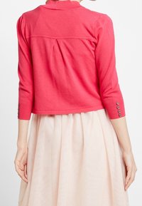 Koraalrode cardigan met korte mouwen en een ronde halslijn, nauwsluitend ontwerp, met drie knoopaccenten op iedere manchet, gecombineerd met een lichte rok.