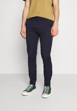 Mężczyzna ubrany w granatowe spodnie slim fit, musztardowy t-shirt i zielone tenisówki z płótna stojący na jednolitym jasnym tle.