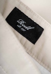 Beige fabric label with black stitching, featuring the word "Reell" in a cursive font and "est. 1997," indicating brand and establishment year.