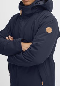 Jūrų mėlynumo pūkinė striukė su aukštu apykakle, užtrauktuku ir odiniu logotipo užtrauktuku. Turinti velcro reguliuojamus rankogalius ir lygią tekstūrą.