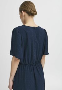 Navy blue dress with short puff sleeves, elastic waistband, and a scooped back featuring six covered buttons. Textured dot pattern throughout.