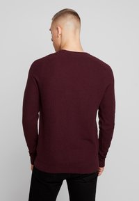 Mann mit kurzen Haaren, der einen burgunderfarbenen Pullover mit langen Ärmeln und schwarze Hosen trägt, steht mit dem Rücken zu einem einfarbigen grauen Hintergrund.