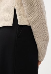 Beiger Strickpullover mit strukturierter Textur und geschlitztem Saum, darüber getragen ein glatter schwarzer Hosenanzug mit Seitentaschen und taillierter Passform.