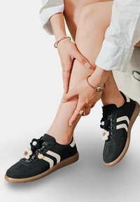 Des baskets en toile noires avec des rayures blanches présentent des pompons décoratifs sur les lacets et une semelle en caoutchouc brun texturé.