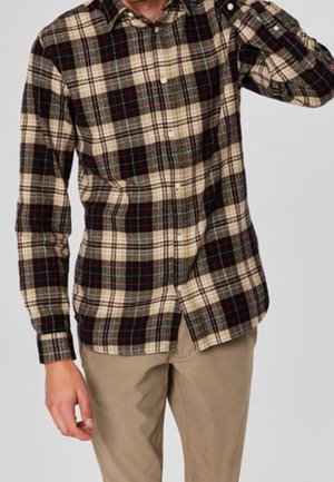 Mann trägt ein beiges, schwarzes und rotes kariertes Hemd mit Knöpfen und Khakihosen, steht vor einem schlichten hellen Hintergrund.