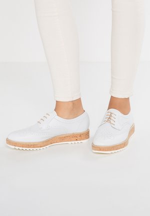 Witte leren schoenen met geperforeerd ontwerp, lichtbruine kurken tussenzool en rubberen zool. Veters in dezelfde kleur als de schoenen. Draagt met lichte broek.