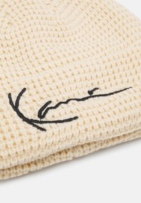 Strickmütze in Creme mit strukturiertem Muster; ausgestattet mit schwarzem, gesticktem Signaturdetail. Weiches Material mit abgerundetem oberen Teil.