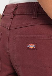 Burgunderfargede bukser med en strukturert stoff, som har baklommer og et Dickies-merkelapp sydd på høyre lomme.