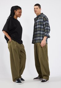 T-shirt noir oversized, pantalon large vert olive. Homme en chemise à carreaux bleu/vert, tous deux en baskets décontractées, sur fond blanc.
