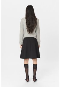 Maglione grigio a maniche lunghe, gonna nera svasata, calze nere a ginocchio con motivo e scarpe nere con tacco. Capelli lisci e scuri. Sfondo minimale.