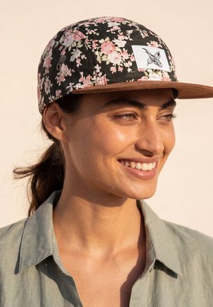 Vrouw die lacht, draagt een zwarte bloemencap met roze bloemen en een bruine klep, gekleed in een lichtgroene blouse met kraag.