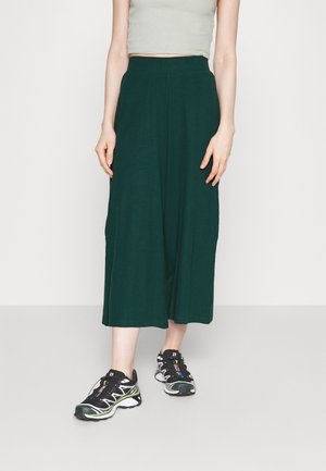 Frau trägt dunkelgrüne, weit geschnittene, knöchellange Hose, helles ärmelloses Oberteil und schwarze Sportschuhe, steht auf einem grauen Boden.