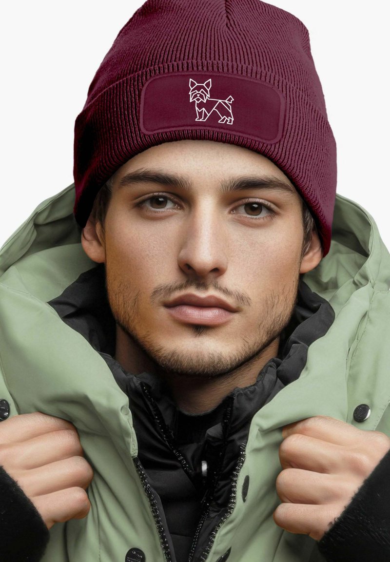 Maroon knitted beanie with a textured pattern and a rectangular patch featuring a white dog silhouette. Worn with a green puffer jacket.