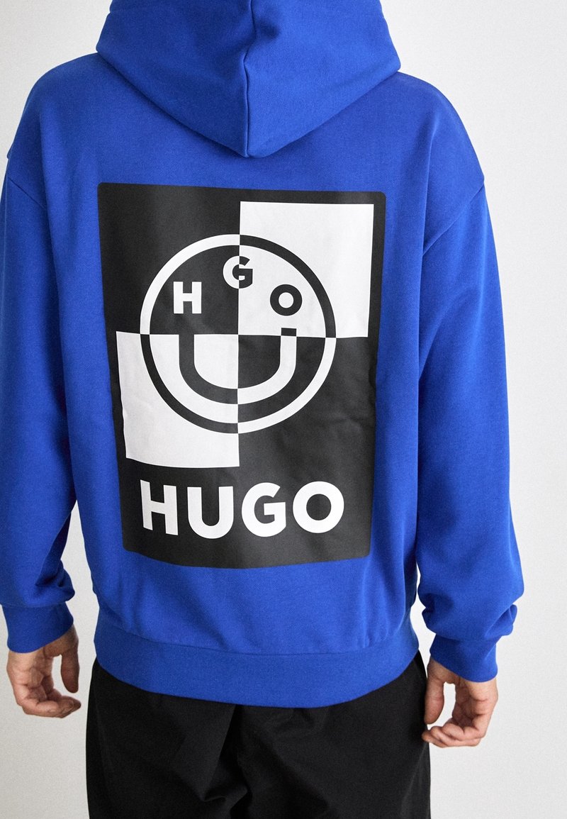 Blå huvtröja med en stor svartvit grafik på baksidan som har en smiley-ansiktsdesign och texten "HUGO" nedanför.