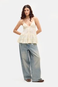 Balta, laisva camisole su giliu V formos iškirpimu ir nėriniu, derinama su plačiomis šviesiai mėlynomis džinsomis. Modelis stovi prieš paprastą foną.