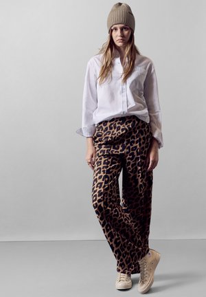Witte button-up shirt, bruine broek met luipaardprint, beige sneakers en een beige gebreide beanie. Relaxte pasvorm met zichtbare stoftexturen.