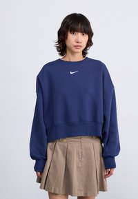 Temno modra skrajšana trenirka z okroglim izrezom in dolgimi rokavi. Ima bel Nike swoosh logotip na prsih. V kombinaciji je z bež plisirano krilo.
