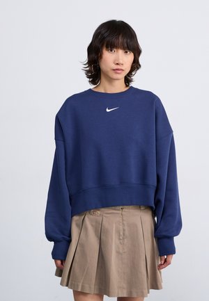 Tumesinine lühike sviiter ümmarguse kaelusega ja pikkade varrukatega. Rindkeres on valge Nike swoosh logo. Koos beeži volditud seelikuga.