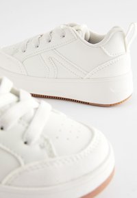 Hvide lædersneakers med rund tå, snørebånd, mesh-foring og en tekstureret gummsoled med en brun accent i bunden.