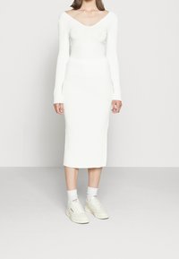 Bílé žebrové pletené šaty s výstřihem do V a dlouhými rukávy, v kombinaci s bílými teniskami a ponožkami do kotníků. Mají přiléhavý střih a délku do poloviny lýtek.