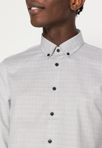 Camicia grigia a scacchi con colletto button-down, bottoni neri e una texture sottile. Presenta un piccolo logo sul lato sinistro.