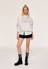 Felpa grigia chiaro con testo stampato, sovrapposta a una camicia bianca con colletto e pantaloni corti neri. Stivaletti neri con lacci e calze grigie.