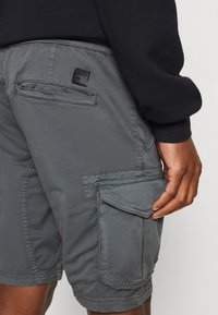 Shorts cargo gris en tissu texturé, avec de multiples poches et un détail cousu. Une petite étiquette noire est visible près de la taille.
