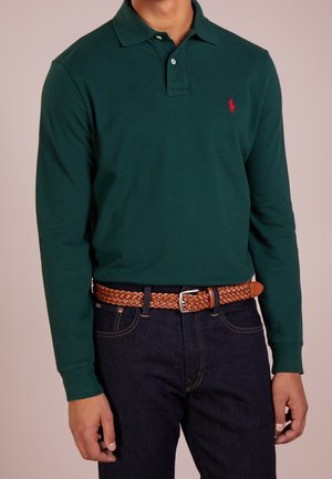 Groen poloshirt met lange mouwen en een knoopsluiting met twee knopen, voorzien van een klein rood logo. Draagt hierbij een donkerblauwe spijkerbroek en een bruine gevlochten leren riem.