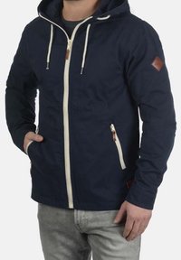 Veste à capuche bleu marine en tissu léger, avec une fermeture éclair frontale, des accents crème et un écusson logo en cuir marron sur la manche.
