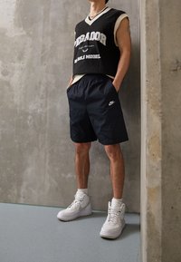 Svart ärmlös tröja med texten "DEGADOR", marinkblå shorts med sidofickor och vita hög-top Nike sneakers. Texturerad betongbakgrund.