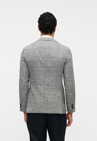 Άνδρας με κοντά σγουρά μαλλιά, ντυμένος με γκρι καρό  σακάκι και μαύρο παντελόνι, στέκεται γυρισμένος με την πλάτη προς ένα απλό λευκό φόντο.