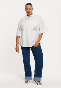 Witte knoop-down shirt met decoratieve zak en korte mouwen, gecombineerd met blauwe wijde jeans en platte sandalen.