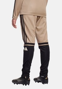 Beige Trainingsanzughose mit schwarzen Akzenten und drei charakteristischen schwarzen Streifen an den Seiten, mit schmaler Passform und Reißverschlussbündchen.