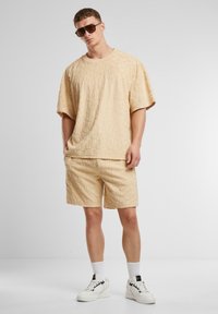 Beige texturovaný top s krátkým rukávem a odpovídající kraťasy. Tkanina má reliéfní vzor. V kombinaci s bílými teniskami a tmavými slunečními brýlemi.