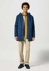 Parka azul marino con capucha, dos bolsillos frontales y cierre de cremallera, usada sobre una camisa beige y pantalones color beige, combinada con zapatos negros.