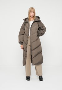 Abrigo acolchado largo en marrón oscuro con capucha, que presenta un patrón de acolchado en forma de flecha, cierre de cremallera y mangas holgadas. Combinado con pantalones beige.