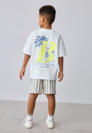 Gyerek fehér pólóban sárga "B" betűvel és kék pálmafa grafikával, csíkos rövidnadrágban és fehér tornacipőben, szürke padlón áll.