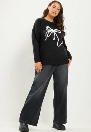 Donna che indossa un maglione nero con design a fiocco bianco, jeans grigio scuro a gamba larga e stivali neri, in piedi davanti a uno sfondo chiaro e uniforme.
