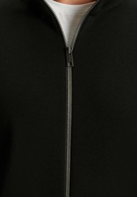 Schwarzer Reißverschluss-Pullover mit glatter Textur, versehen mit einem silbernen Reißverschluss und einem hohen Kragen. Darunter ist der Halsausschnitt eines weißen T-Shirts.