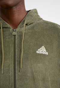 Olivgrön, kordfloddszip-upp hoodie med en fodrad textur, dragskor och vitt Adidas-logotyp på vänster bröst.