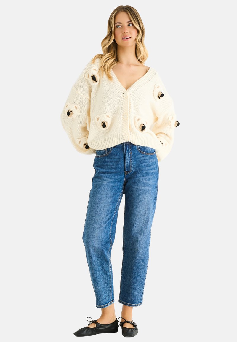 Cream knitted cardigan featuring teddy bear appliqués, V-neck design, and button closure, paired with high-waisted blue jeans and black flats.