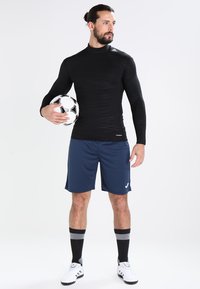Sort langærmet atletisk top med struktureret mønster, marineblå shorts, sort og hvid fodbold, hvide og sorte sportssko med stribede sokker.