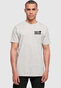 Lysegrå bomulds t-shirt med korte ærmer og rund halsudskæring. Har sort trykt tekst på venstre brystområde. Renssym, enkelt design.