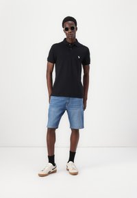 Črna polo majica z majhnim belim logotipom, v kombinaciji z modrimi denim kratkimi hlačami. Model nosi sončna očala in bež superge z črnimi nogavicami.