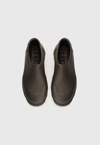 Sötétbarna slip-on cipők sima, matt felülettel, ívelt formával és diszkrét részletezéssel a felső részen. A belső talp kontrasztos rózsaszín szöveget mutat.