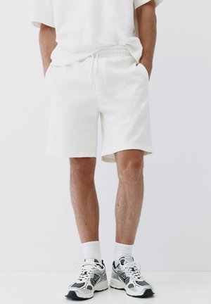 Homme portant un short décontracté blanc, des chaussettes blanches et des chaussures de course noires, blanches et grises sur un fond uni.