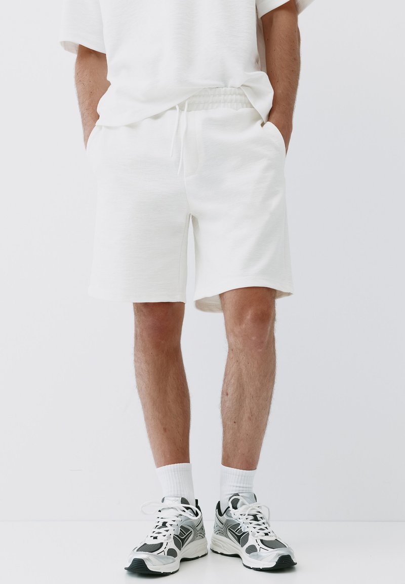 Homme portant un short décontracté blanc, des chaussettes blanches et des chaussures de course noires, blanches et grises sur un fond uni.