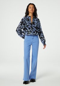 Schwarze Bluse mit blauem und grünem Blumenmuster, geknöpftem Ausschnitt und voluminösen Ärmeln, kombiniert mit hellblauen, weit geschnittenen Jeans und schwarzen Schuhen.