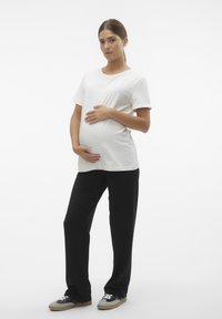 T-shirt de algodão branco com mangas curtas, combinado com calças largas pretas, usado por uma modelo grávida que segura a sua barriga. Calçado casual incluído.
